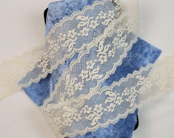May include: Ivory floral lace ribbon with a scalloped edge, displayed on blue velvet fabric. The lace features a delicate floral pattern, ideal for adding elegance to craft projects or clothing.