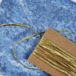 Könnte beinhalten: Nahaufnahme eines goldenen Bandes auf einem braunen Kartonträger, mit einem blauen Samtstoff im Hintergrund. Das Band ist dünn und hat ein strukturiertes, metallisches Aussehen. Der Stoff hat eine weiche, plüschige Textur.