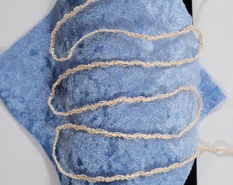 Puede incluir: Una tela de terciopelo azul claro con un ribete decorativo color crema. La tela está doblada y en capas, con el ribete creando un patrón ondulado. Un objeto negro es parcialmente visible detrás de la tela.
