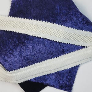 Puede incluir: Primer plano de tela y adornos. Un adorno de color crema con borde festoneado y centro tejido se coloca sobre un trozo de tela de terciopelo azul oscuro. Un trozo de tela negra es visible debajo.