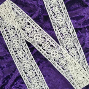 May include: Ivory-colored lace ribbon with a floral and geometric pattern. The ribbon is laid out on a deep purple velvet surface. The lace has a delicate, openwork design, perfect for crafting or adding a vintage touch to projects.