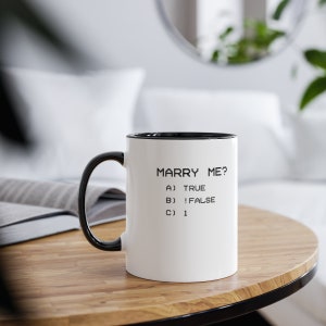 May include: White ceramic mug with a black interior, handle, and rim. The mug features the text "MARRY ME?" followed by multiple-choice answers: A) TRUE, B) !FALSE, and C) 1. The mug is on a wooden table.