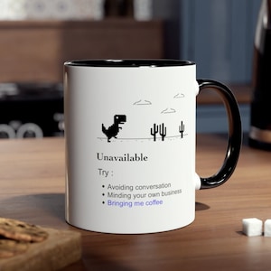 Pode incluir: Caneca de cerâmica branca com interior e alça pretos. A caneca apresenta um dinossauro pixelado com o texto "Unavailable" e uma lista de sugestões humorísticas, incluindo "Bringing me coffee."