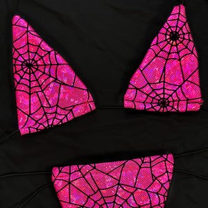 May include: A two-piece swimsuit set in bright pink, featuring a spiderweb design in black. The bikini top has triangle cups, and the bottoms are a classic cut. The set is displayed on a black background.
