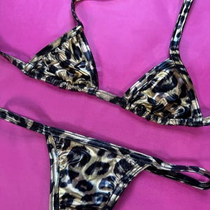 Puede incluir: Un conjunto de bikini de dos piezas con estampado de leopardo. La parte superior es de estilo triangular con tirantes halter, y la parte inferior es de estilo tanga. La tela tiene un acabado brillante y metálico con manchas negras y doradas. El bikini se muestra sobre un fondo rosa.