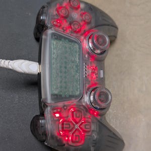 May include: A transparent gaming controller with red illuminated buttons and a small screen. The controller is connected to a white cable and rests on a wooden surface. The buttons include a directional pad and action buttons.