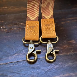 May include: Two dog leash straps with a camouflage pattern in shades of brown and tan. The straps have brown leather accents with the "SSO" logo and brass-colored swivel clasps.