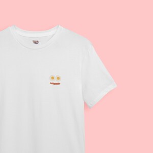 Könnte beinhalten: Weißes T-Shirt mit einem Aufdruck von zwei Spiegeleiern und einer Scheibe Speck.
