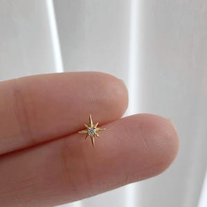 Puede incluir: Un pequeño pendiente dorado en forma de estrella con una sola piedra preciosa clara y pequeña en el centro. El pendiente se sostiene entre dos dedos sobre un fondo blanco.