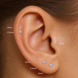 Puede incluir: Primer plano de una oreja con varios pendientes de botón de ópalo de diferentes tamaños. Los pendientes tienen un diámetro de 2 mm, 2,5 mm y 3 mm.
