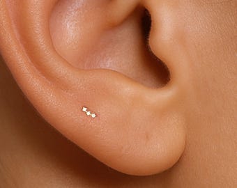 Puede incluir: Un pequeño pendiente de botón en oro con tres diminutas piedras claras de corte redondo dispuestas en una línea horizontal recta. El pendiente está colocado en el lóbulo de la oreja, mostrando un diseño delicado con un acabado de metal pulido.