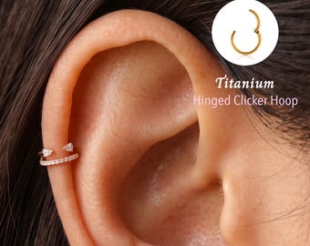May include: A gold-toned hinged clicker hoop earring featuring a row of small clear stones and two triangular-cut stones at the ends. The text Titanium Hinged Clicker Hoop is displayed next to a circular inset showing the hoop in an open position.