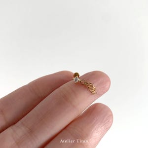 K&ouml;nnte beinhalten: Ein zarter goldener Ohrring mit einem kleinen, klaren Edelstein und einem h&auml;ngenden Kettendesign. Der Ohrring wird zwischen den Fingern vor einem wei&szlig;en Hintergrund gehalten. Der Text "Atelier Titan" ist unten sichtbar.