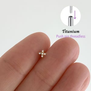 May include: A small, gold-colored, cross-shaped earring with four clear gems and small white pearls. The text "Titanium Push pin threadless" is visible in the image, along with a diagram of the earring's post.