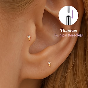 Puede incluir: Pendientes de botón dorados con una pequeña gema transparente en la parte superior. Los pendientes tienen forma de pequeña llama estilizada. El texto "Titanium Push pin threadless" es visible en la imagen.