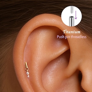 May include: A gold-colored, threadless push pin earring with two small, clear gemstones. The earring is shown in an ear piercing. The image also shows a diagram of the earring's push pin mechanism. The text "Titanium Push pin threadless" is also visible.