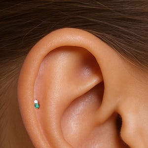 May include: A close-up of a person's ear with a small emerald green and white gemstone stud earring.