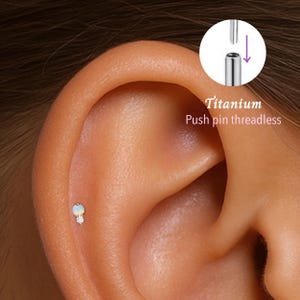 May include: A close-up of an ear with a small, gold-colored earring featuring a white opal and a clear crystal. The earring is a push-pin, threadless design made of titanium. The text "Titanium Push pin threadless" is visible.