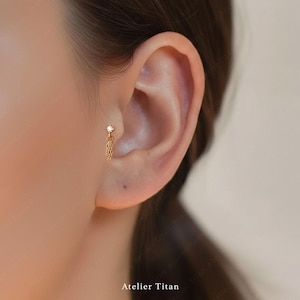 K&ouml;nnte beinhalten: Goldfarbener Ohrring mit einem kleinen, klaren Edelstein und einem h&auml;ngenden Ketten-Design. Der Ohrring wird am Ohr getragen. Der Text "Atelier Titan" befindet sich am unteren Rand des Bildes.