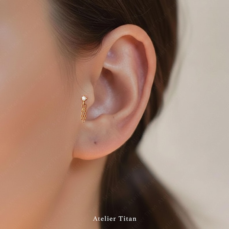 K&ouml;nnte beinhalten: Goldfarbener Ohrring mit einem kleinen, klaren Edelstein und einem h&auml;ngenden Ketten-Design. Der Ohrring wird am Ohr getragen. Der Text "Atelier Titan" befindet sich am unteren Rand des Bildes.