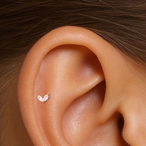 May include: A gold-colored ear piercing with two clear, marquise-cut gemstones. The jewelry is set on the upper ear cartilage, showcasing a delicate and elegant design. The piercing is small and subtle, suitable for everyday wear.