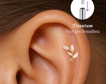 May include: A gold-colored ear piercing with a leaf design, featuring clear, marquise-cut stones. The text "Titanium Push pin threadless" is visible. The piercing is shown on an ear.