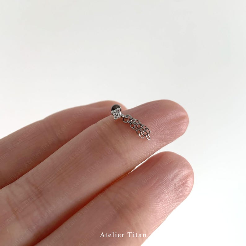 K&ouml;nnte beinhalten: Ein kleiner, silberfarbener Ohrring mit einem h&auml;ngenden Ketten-Design. Der Ohrring hat einen kleinen, schwarzen Stein an der Spitze, umgeben von klaren Steinen. Eine Kette h&auml;ngt darunter und verleiht dem Schmuck eine zarte Note.