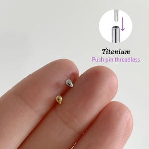 Może przedstawiać: Zbliżenie dwóch kolczyków do ciała w kształcie łezki. Jeden jest złoty, drugi srebrny. Widoczny jest tekst "Titanium Push pin threadless".