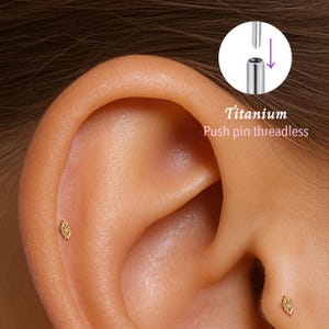 May include: Close-up of an ear with a small, gold-coloured stud earring. The earring has a decorative, floral design. The image also shows a diagram of a titanium push pin threadless earring.