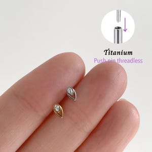Pode incluir: Dois brincos de titânio sem rosca em forma de gota. Um brinco é dourado, o outro prateado, cada um com uma gema transparente no centro. A imagem inclui um diagrama mostrando como os brincos sem rosca de pino funcionam. O texto diz "Titanium Push pin threadless."