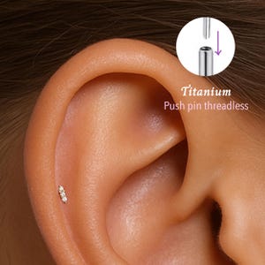 Könnte beinhalten: Nahaufnahme eines Ohrs mit einem kleinen, goldfarbenen Ohrring mit drei kleinen, klaren Edelsteinen. Der Ohrring ist ein Push-Pin-Design ohne Gewinde. Der Text lautet "Titanium".