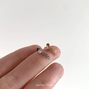 K&ouml;nnte beinhalten: Zwei zarte Ohrringe mit Kettenakzenten. Ein Ohrring ist silberfarben mit einem dunklen Edelstein und einer Kette, der andere ist goldfarben mit einem klaren Edelstein und einer Kette. Die Ohrringe sind klein und elegant.