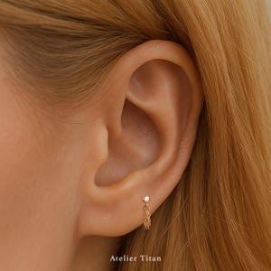 K&ouml;nnte beinhalten: Nahaufnahme eines Ohrs mit einem kleinen goldenen Ohrring. Der Ohrring hat einen einzelnen, klaren Edelstein-Stecker und einen zarten Kettenanh&auml;nger. Der Text "Atelier Titan" befindet sich am unteren Bildrand.
