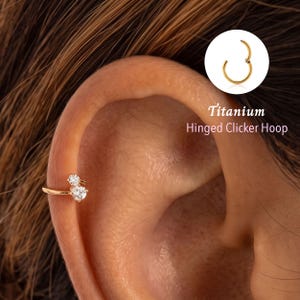 Pode incluir: Um brinco de argola com fecho de clique em tom dourado, apresentando duas pedras transparentes de corte redondo dispostas verticalmente. A joia é exibida numa orelha, com um detalhe mostrando o design da argola e o texto Titanium Hinged Clicker Hoop.