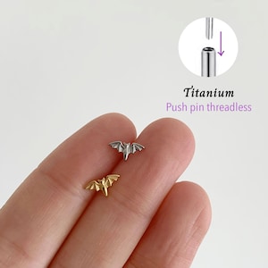 Könnte beinhalten: Zwei kleine fledermausförmige Körperschmuckstücke, eines silberfarben und eines goldfarben, werden gezeigt. Der Schmuck besteht aus Titan und hat ein gewindefreies Push-Pin-Design. Der Text "Titanium Push pin threadless" ist ebenfalls sichtbar.