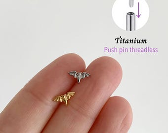 May include: Two small bat-shaped body jewelry pieces, one silver and one gold, are displayed. The jewelry is made of titanium and has a push pin threadless design. The text "Titanium Push pin threadless" is also visible.