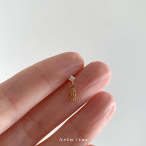 K&ouml;nnte beinhalten: Goldfarbener Ohrring mit einem kleinen, klaren Edelstein und einer herabh&auml;ngenden Kette. Der Ohrring wird zwischen Fingern gehalten. Der Text "Atelier Titan" ist unten sichtbar.