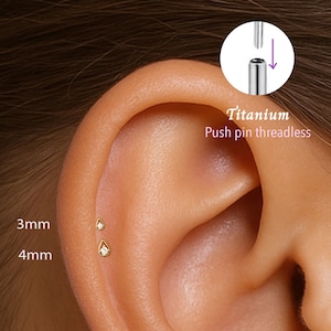 May include: An ear with two gold-coloured, teardrop-shaped earrings, 3mm and 4mm in size, each with a small, clear gem. The text "Titanium Push pin threadless" is visible. The earrings are shown in situ.