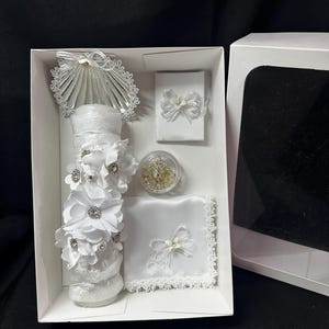 May include: A white gift box containing a decorated candle, a small box with a bow, a small container of beads, and a handkerchief with a bow. The candle is adorned with white flowers and rhinestones.