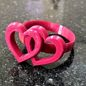 May include: A magenta-colored, interlocking heart-shaped ring. The ring features two connected hearts, with a band that wraps around the back. The design is likely 3D-printed, with visible layer lines. The ring is a symbol of love and commitment.