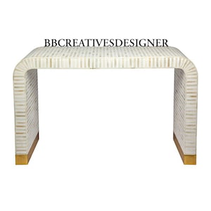 Puede incluir: Una mesa consola de incrustaciones de hueso blanco con base dorada. La mesa tiene una tapa rectangular y un diseño curvo en las patas.