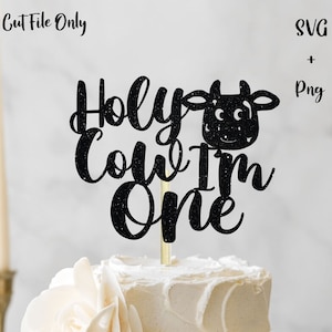 Puede incluir: Topper de pastel negro con purpurina con el texto "Holy Cow I'm One" y una cabeza de vaca con ojos blancos. El topper está en un pastel blanco con glaseado blanco.