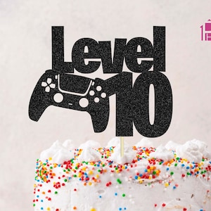 Puede incluir: Topper de pastel negro con purpurina con las palabras "Level 10" y un mando de videojuego. El topper de pastel está sobre un pastel blanco con chispas de colores.
