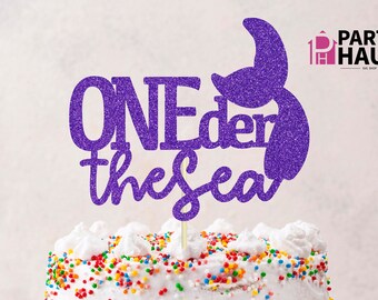Pode incluir: Topper de bolo de purpurina roxa com o texto "ONEder the sea" e uma cauda de sereia.