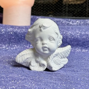 May include: White ceramic angel figurine with a cherub face and feathery wings. The angel is sitting on a purple sparkly surface.