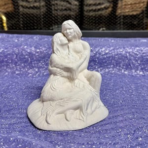 May include: A white ceramic sculpture depicting two figures embracing. One figure is seated, and the other is standing, with their arms wrapped around each other. The sculpture has a smooth, matte finish and is set against a purple background.