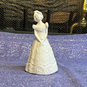 May include: White porcelain figurine of a woman in a long, lace dress. The figurine is standing on a purple fabric background.