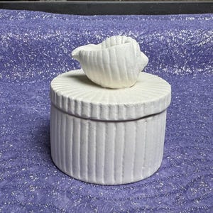 May include: White ceramic trinket box with a seashell lid. The box has a ribbed texture and the lid is shaped like a seashell.