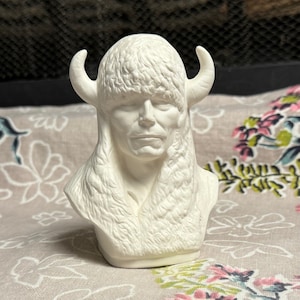 May include: A white ceramic bust of a person wearing a horned headdress and fur-lined hood. The sculpture has a detailed face and is set against a floral patterned background.