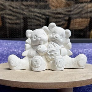 May include: Two white ceramic teddy bears holding a baseball and a glove. The bears are sitting on a wooden base.